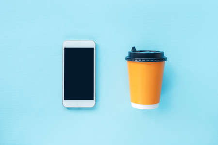 Coffee in yellow paper cup and cell phone on blue background. Food delivery and takeout concept. Top view, flat lay.の写真素材