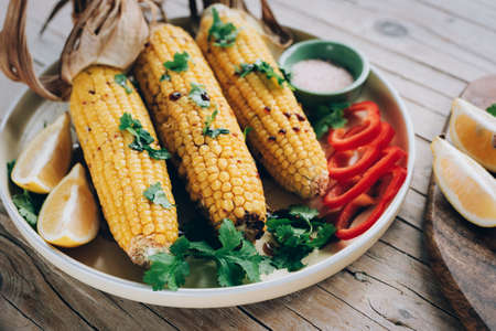 Baked corn cobs with sauce, coriander, sliced bell pepper, lemon and salt on wooden table. Mexican food. Top view.の写真素材