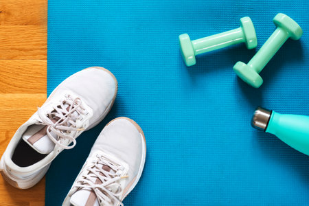 Light green dumbbells, bottle with water and white sneakers on blue sports mat background. Training...