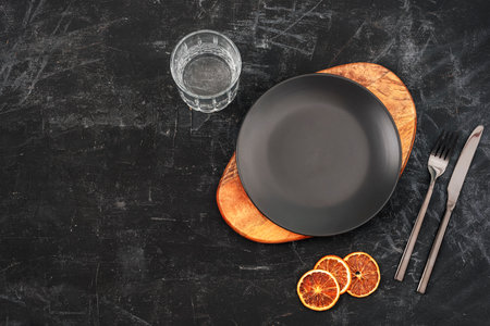Black table setting with a plate on cutting board, glass of water, fork and knife and orange slices. Top view, flat lay, copy spaceの写真素材