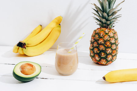 Still life with fruits, banana, pineapple and avocado smoothie on white wooden table with long shadows. Minimal detox diet concept, summer vitamin drink.の写真素材