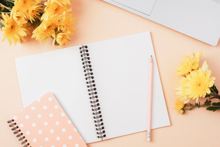 Desktop with notebook, laptop computer, pencil, bouquet of yellow flowers on beige background. Top view, flat lay.の写真素材