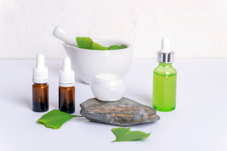 Fresh green ginkgo leaves, cream jar on a natural stone, brown glass bottles with essential oil, face serum bottle and mortar and pestle on white background. Modern apothecary, natural ingredientsの写真素材
