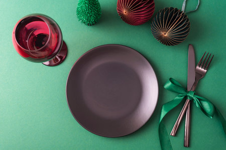 Festive table setting with black plate, fork and knife, wineglass and Christmas decorations on green background. Top view, flat lay, copy spaceの写真素材
