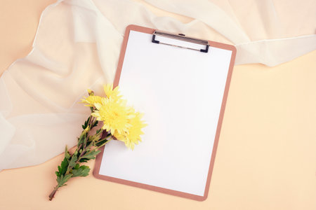Clipboard with blank paper and yellow chrysanthemums on beige background. Top view, flat lay,...