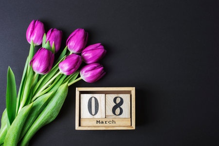 Bouquet of purple tulips and wooden block calendar with 8 march date on black table. Womens day...