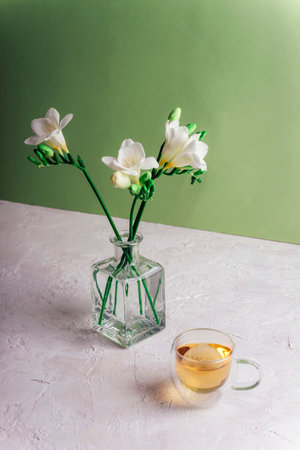 A cup of tea and freesia white flowers in glass vase on white textured table against green wall. Still life.の写真素材