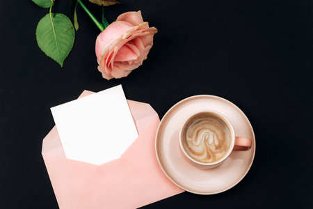 A coffee cup, pink rose and envelope with blank card on black background. Holiday concept. Top view, flat lay, mockup.の写真素材