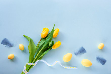 Easter festive background. Yellow painted eggs, feathers and tulips on blue table. Top view, flat lay, copy spaceの写真素材