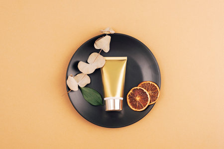 Golden cream tube and dry orange slices, eucalyptus leaves on black plate on beige background. Top view, flat lay, mockup.の写真素材