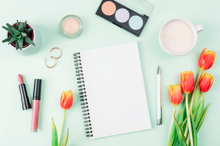 Blank notepad, cup of coffee, make up cosmetics and bouquet of orange tulips on light green background, top view. Womens day or Mothers day concept. Creative flat lay, mockupの写真素材