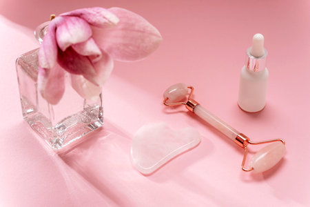 Rose quartz crystal facial roller and gua sha scraper, face serum and magnolia flower in vase on pink background. Facial massage kit for lifting therapy . Skin care anti-aging tools. Top view.の写真素材