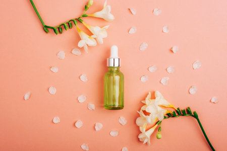 Green cosmetic serum bottle and white freesia flowers on coral pink background. Top view, flat lay, mockup.の写真素材