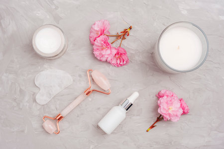 Cream jar, face roller, gua sha stone and pink cherry blossom on gray concrete background. Skin care, beauty treatment concept. Top view, flat lay.の写真素材