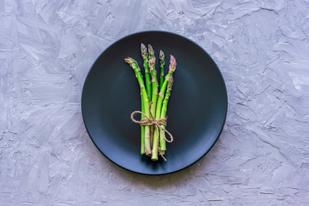 Fresh green asparagus on black plate on grey concrete background. Healthy vegan raw food concept. Top view, flat layの写真素材