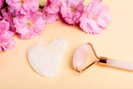 Face roller, gua sha stone and pink cherry blossom on beige background. Skin care, beauty treatment concept. Top viewの写真素材