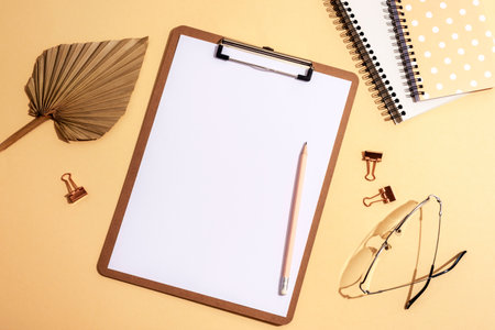 Desktop with blank clipboard, notepads, fan leaf, eyeglasses and accessories on neutral beige background. Top view, flat lay, mockup.の写真素材