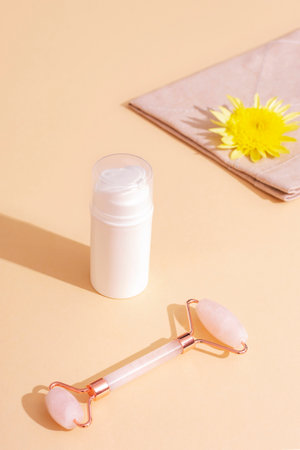White cosmetic bottle, yellow flower and rose quartz facial roller on beige background in sunlight. Skincare, spa and wellness concept.の写真素材