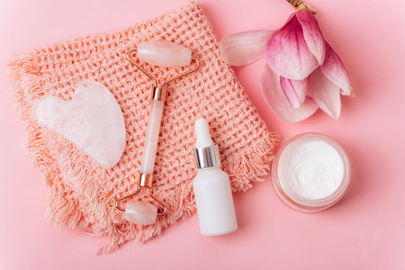 Rose quartz crystal facial roller and gua sha scraper, face serum or organic cosmetic oil, cream jar and magnolia flower on pink background. Facial massage kit for lifting therapy. Top view, flat lay.の写真素材