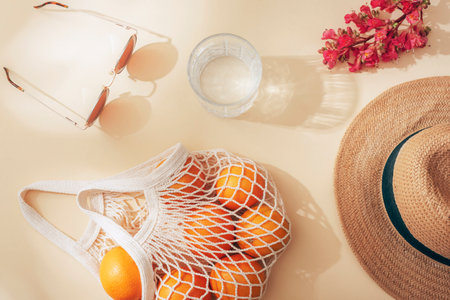 Straw hat, sunglasses, glass of water, string bag with oranges and red flower on beige background in...