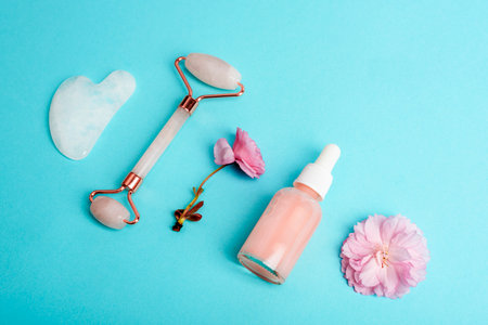 Serum bottle, face roller, gua sha stone and pink sakura blossom on turquoise blue background. Skin care, beauty treatment concept. Top view.の写真素材