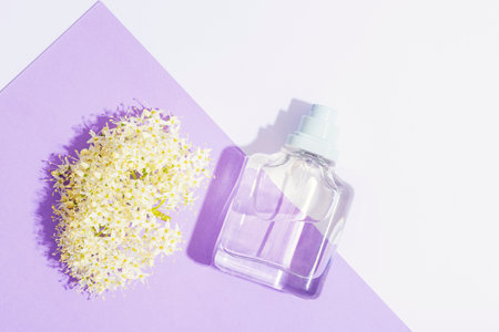 Glass perfume bottle and white blossom on white and lilac background in sunlight. Top view, flat...