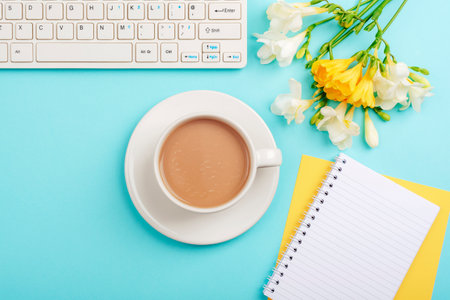 Computer keyboard, blank notepad, pen and coffee cup on blue table with yellow freesia flowers. Top view, flat lay, mockupの写真素材