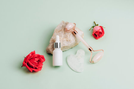 White cosmetic serum bottle, face roller, gua sha stone and red rose flowers on natural stone on light green background. Skin care, beauty treatment concept. Top viewの写真素材