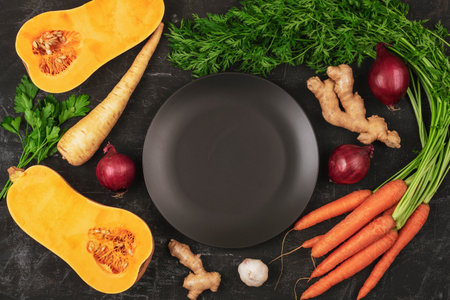 Black plate and vegetables, pumpkin. carrot, ginger, onion, parsnip and parsley on black background. Healthy raw vegan food. Top view, flat lay, mockup.の写真素材