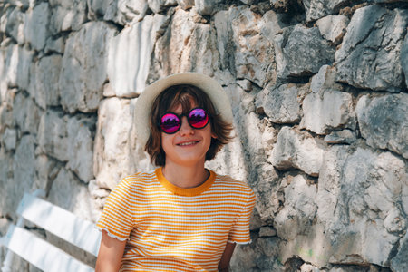Beautiful happy girl in straw hat and sunglasses sitting on the bench against stone wallの写真素材