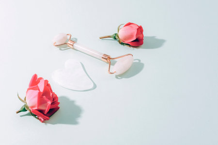 Face roller, gua sha stone and pink rose flowers on light green background with harsh shadows. Skin care, beauty treatment concept. Top view, flat lay, copy space.の写真素材