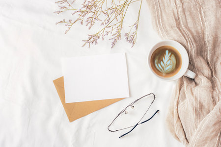 Coffee cup, envelope with blank card, glasses and small flowers on white linen. Cozy workplace,...