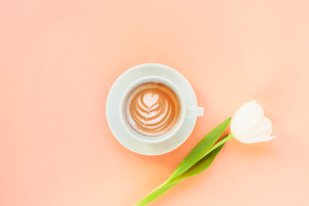 Cup of coffee with coffee art and white tulip on peach color background. Womens day or Mothers day concept. Top view, flat lay, copy spaceの写真素材