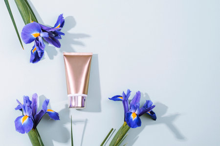 Golden cosmetic cream tube and blue iris flowers on light blue background in sunlight. Top view, flat lay, copy spaceの写真素材