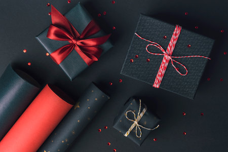 Gift boxes packaging with red bow and rolls of wrapping paper on black background. Festive Christmas, New Year holidays celebration concept. Top view, flat lay.の写真素材