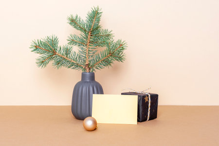 Spruce branch in ceramic vase, gift box and blank card on beige table. Minimal Christmas concept. Front view, mockup.の写真素材