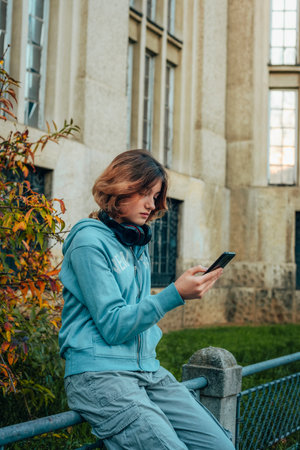 Beautiful teenage girl with a smartphone sits on a fence against the background of a building.の写真素材