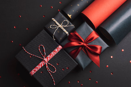 Gift boxes packaging with red bow and rolls of wrapping paper on black background. Festive Christmas, New Year holidays concept. Top view, flat layの写真素材