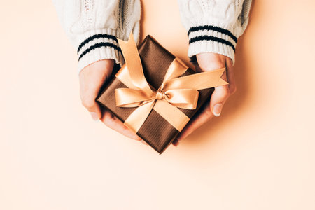 Girls hands in white sweater holding brown gift box with golden bow on light peachy background. Christmas, Valentines day, Black friday sale concept. Top view, flat lay. Peach Fuzz color of the year 2024の写真素材