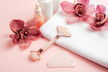 Facial cosmetic roller and gua sha scraper with pink orchid flowers on a white towel. Home spa concept.の写真素材