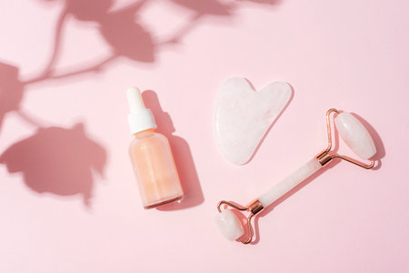 Cosmetic serum bottle, face roller and gua sha stone with orchid flower shadow on pink background. Sharp shadows. Home spa concept. Top view, flat lay.の写真素材