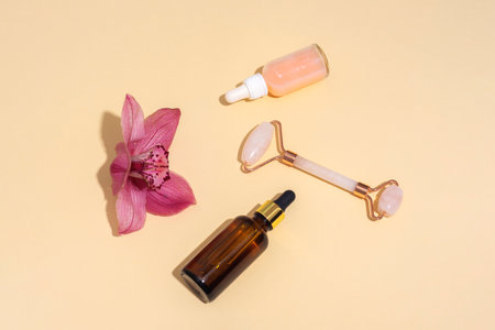 Cosmetic bottles and face roller with pink orchid flower on beige background in sunlight. Skin care, spa and wellness concept. Top view, flat lay.の写真素材