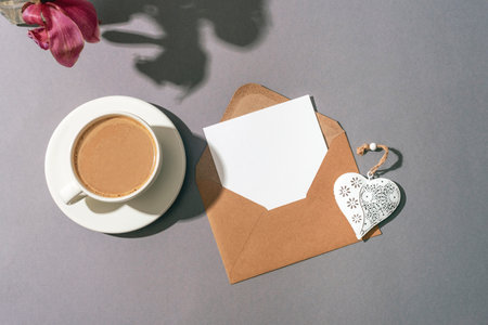 Envelope with blank card, white vintage heart, coffee cup and flowers shadow on gray background. Top view, flat lay, mockup.の写真素材
