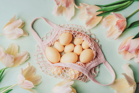 String bag with Easter eggs and yellow tulips on light green background. Top view, flat lay.
