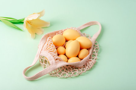 String bag with Easter eggs and yellow tulip on light green background. Top view, flat lay.