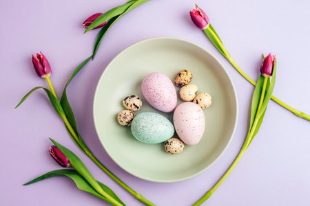 Easter table setting with green plate, purple tulips and Easter eggs on lilac background. Top view,...