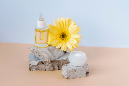 Cosmetic serum bottle and cream jar on stone podium with yellow gerbera flower on beige table. Closeup.の写真素材