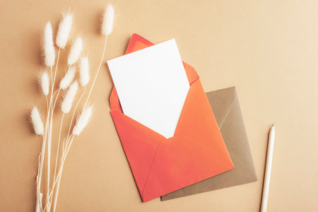 Red envelope with blank card, pencil and Lagurus Ovatus flowers on brown background. Top view, flat...