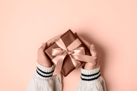 Girls hands in white sweater holding brown gift box with golden bow on neutral background. Top view, flat lay.の写真素材
