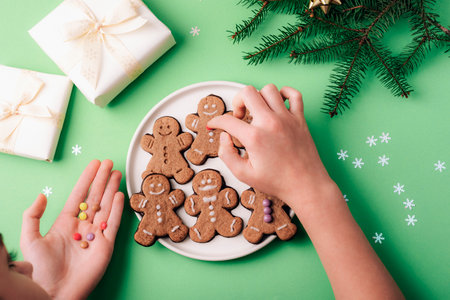 Top view of hands decorating gingerbread cookies with festive decorations, wrapped gifts, and pine branches on a green background.の写真素材